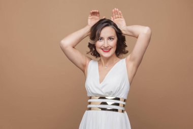 Tavşan kulağı gösterilen ve kahverengi arka plan üzerinde gülümseyen beyaz elbiseli komik orta yaşlı kadın