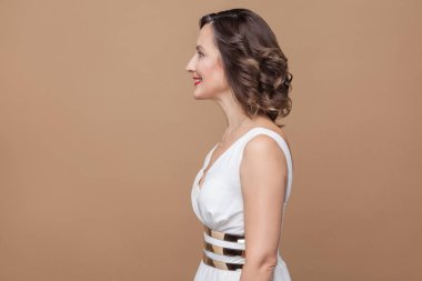 Dişlek gülümsüyor mutlu orta yaşlı kadın kırmızı dudaklar ve kahverengi arka plan üzerinde poz karanlık kıvırcık saç modeli ile beyaz elbiseli yan görünüm