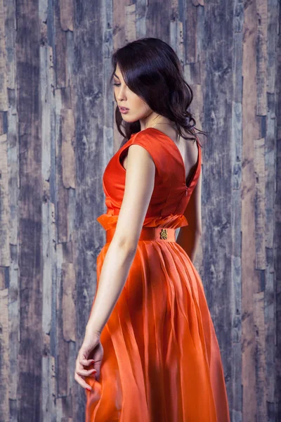 年轻的黑发妇女穿着时髦的橙色丝绸礼服摆在木质背景上 — 图库照片