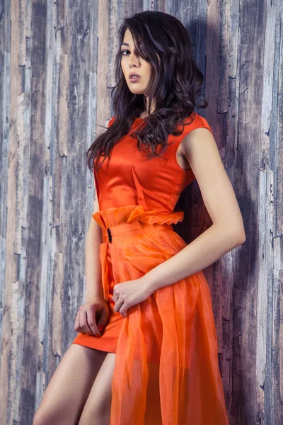 年轻的黑发妇女穿着时髦的橙色丝绸礼服摆在木质背景上 — 图库照片