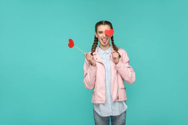 Ayakta ve kırmızı kalp çıkartmaları yeşil arka plan üzerinde dilini çıkarmış kameraya bakarken tutan helezon saç modeli ile rahat tarzı komik güzel kadın 