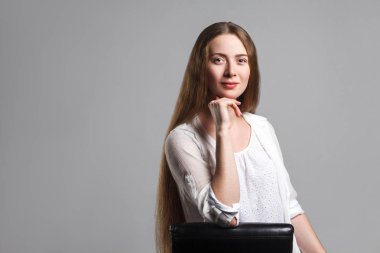 Siyah sandalyede oturan ve çenesinin dokunmaktan ve gri arka plan üzerinde kameraya bakarak beyaz t-shirt uzun kahverengi saçlı güzel mutlu model portresi