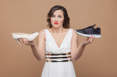 Şüpheli düşünme iş kadını rahat spor ayakkabı ile zarif yüksek topuk tutarak ve kameraya kahverengi zemin üzerinde seyir beyaz elbiseli
