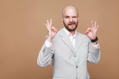 Portrait of handsome funny middle aged bald bearded businessman in light gray suit showing Ok sign and winking on brown background clipart