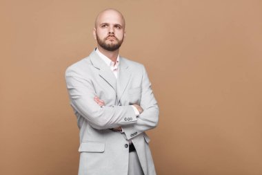 Karışık yakışıklı orta yaşlı kel sakallı işadamı kahverengi zemin üzerinde çapraz elleriyle hafif gri takım ayakta portresi