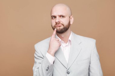 karışık düşünceli yakışıklı orta yaşlı kel işadamı ışık gri elbiseli ayakta ve parmağını çeneme kahverengi zemin üzerinde düşünme sakallı