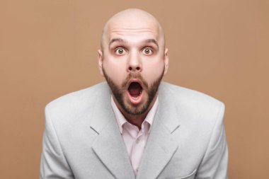 Orta yaşlı kel yakışıklı işadamı kameraya hayran yüz ve açık ağız ile seyir üzerinde kahverengi zemin ışık gri takım ayakta sakallı