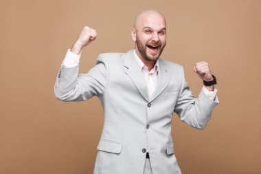Portresi memnun mutlu kazanan orta yaşlı kel sakallı işadamı açık gri takım elbise kahverengi arka plan ayakta ve kutluyor zafer