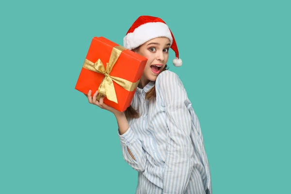 wondered beautiful woman in striped blue shirt in red christmas cap holding red gift box and looking at camera with amazed face on green background, New year concept