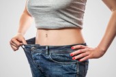 Detail slim pasu mladé ženy ve velkých džínách a šedé nahoře ukazuje úspěšné hubnutí na světle šedém pozadí, dietního konceptu 