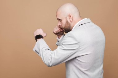 Orta yaşlı kel yan görünüm işadamı ayakta ve kahverengi zemin üzerinde boksör poz ile saldırmaya hazır ışık gri elbiseli sakallı