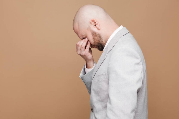 side view of sad crying middle aged bald bearded businessman in light gray suit standing and closed eyes with hand on brown background