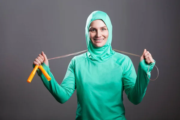 Happy beautiful woman in green islamic sportswear holding jump rope and looking at camera with toothy smile on grey background