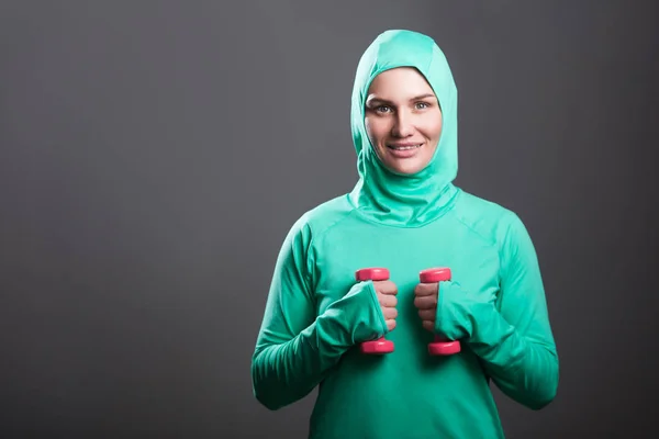 Happy beautiful woman in green islamic sportswear holding red dumbbells in hands and  looking at camera with toothy smile on grey background