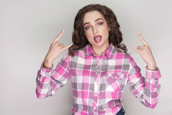 funny beautiful woman with curly hairstyle and makeup in pink checkered shirt looking at camera and showing rock sign with tongue out on gray background