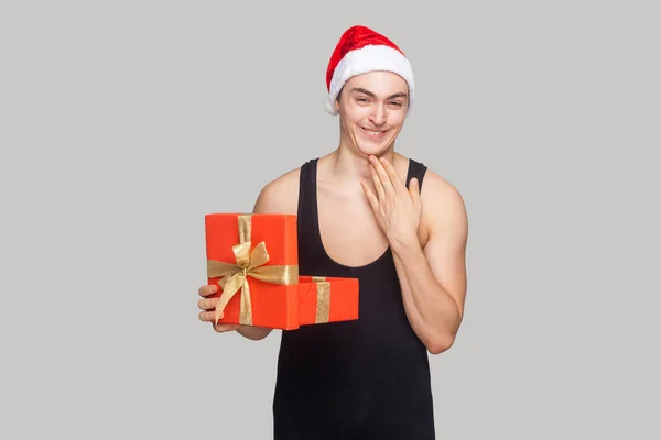 Cunning man in red christmas hat holding gift box and touching his chin while toothy smiling and looking at camera with funny face on gray background