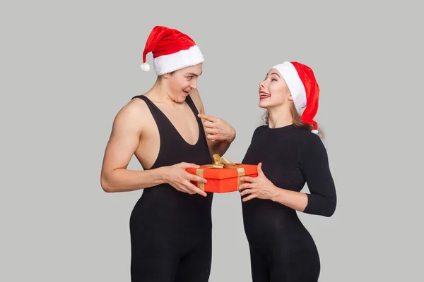 Woman presenting gift box for surprised man in santa hat, on gray background, New year concept