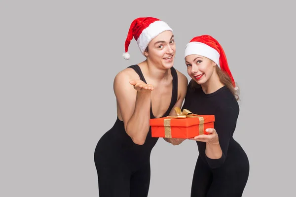 Couple Christmas Hats Presenting Gift Box Looking Camera Gray Background — Stock Photo, Image