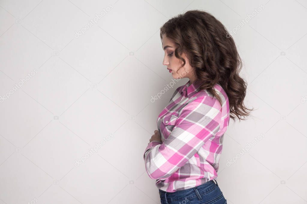 side view of unhappy sad woman with curly hairstyle and makeup in pink checkered shirt standing with crossed hands and looking head down on gray background
