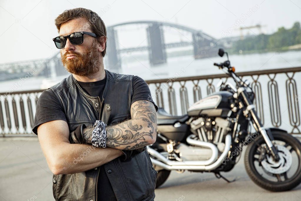 Redhead biker with beard in leather jacket standing with crossed arms near motorbike on bridge 