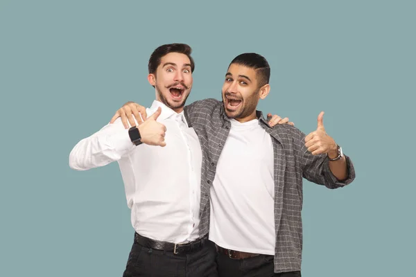 两个英俊的胡子快乐的合作伙伴拥抱和看在相机与满意的惊讶的脸 而在蓝色背景上显示大拇指 — 图库照片