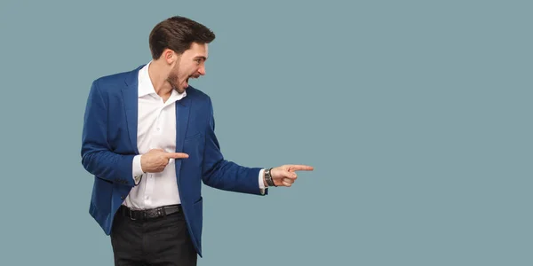 Handsome amazed bearded man in suit pointing aside and screaming on blue background