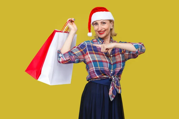 ショッピング バッグ黄色の背景に赤のサンタ キャップ ポインティング指の中年の女性 — ストック写真