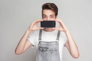 Denim tulum ve holding ve gri arka plan üzerinde kameraya bakarken smartphone ile ağız kapsayan beyaz T-shirt komik modern genç çocuk