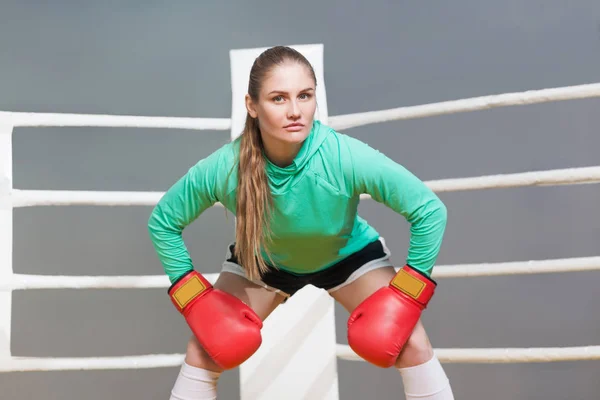 Angry boxing athletic young woman in green long sleeve poising with red boxing gloves and looking at camera with serious face on gray background