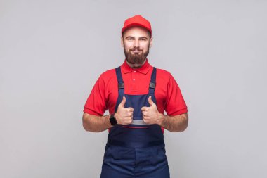 Mavi genel sakallı genç memnun neşeli tamirci ve ayakta ve gri arka plan üzerinde gülümsemeyle thumps gösterilmesini kap ile kırmızı t-shirt portre