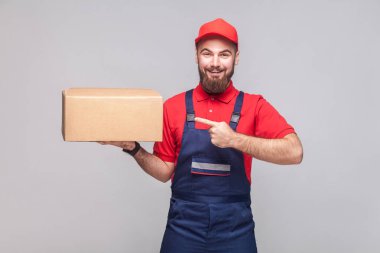 Holding ve gri arka plan üzerine karton kutu, parmakla mavi üniforma ve kırmızı t-shirt sakallı genç neşeli lojistik teslimat adam 