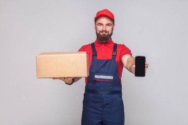 Karton kutu tutarak ve smartphone ekran gri arka plan üzerinde gösterilen mavi üniforma ve kırmızı t-shirt sakallı lojistik adam gülümseyen genç