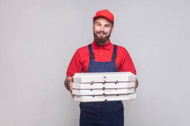 Karton pizza kutuları yığını tutan ve gri arka plan üzerinde kameraya bakarak mavi üniforma ve kırmızı t-shirt sakallı lojistik teslimat adam gülümseyen genç