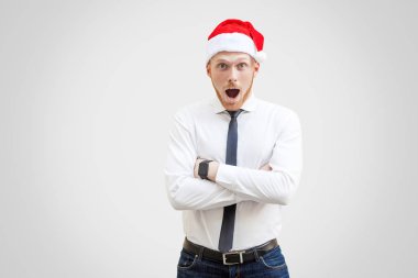 Hayret suratla kameraya bakarak ve ışık gri arka plan üzerinde çapraz eller ile ayakta siyah kravat ve kırmızı yılbaşı şapka ile beyaz gömlekli merak yakışıklı işadamı portresi