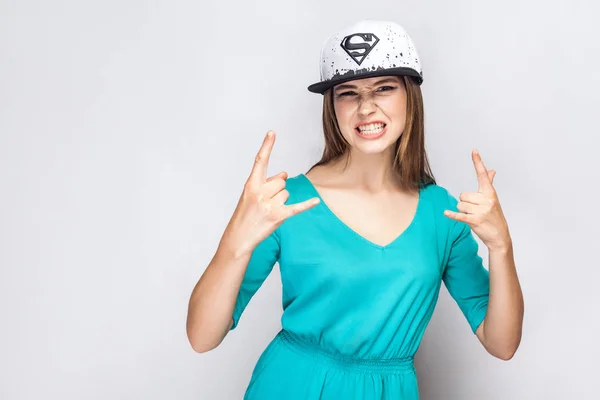 Portrait of aggressive beautiful young woman in blue dress and white cap with superman symbol standing with raised arms and showing rock sign on grey background