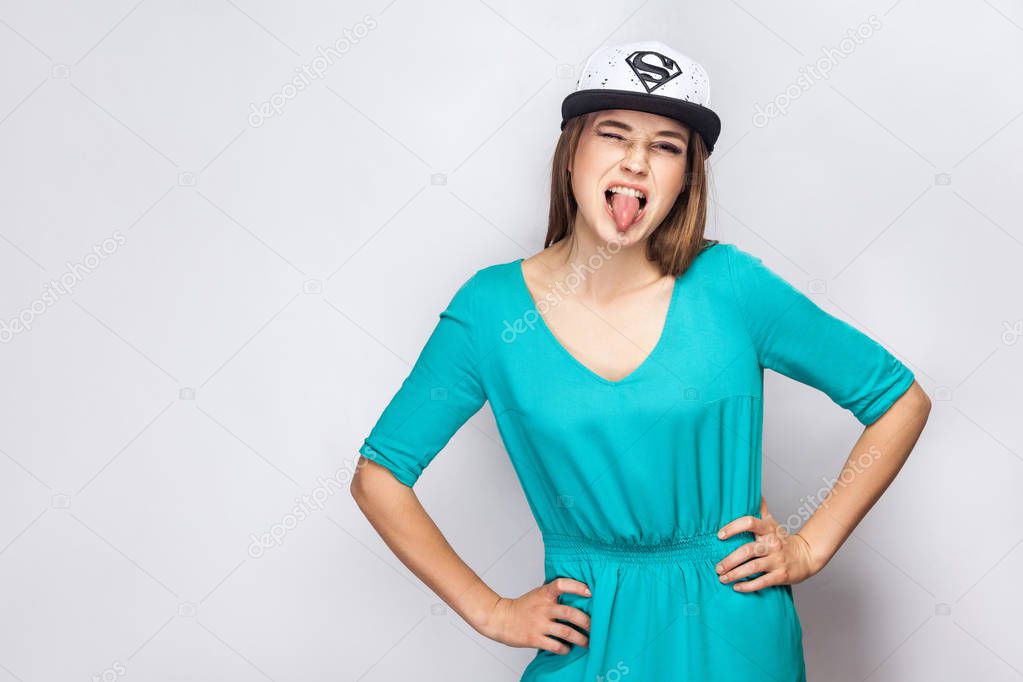 Portrait of crazy beautiful young woman in blue dress and white cap with supermen sign holding hands on waist and winking with tongue out on grey background