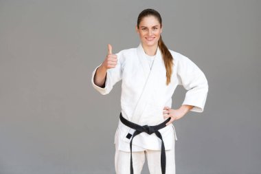 Positive athletic young woman in white kimono and black belt showing thumbs up and looking at camera with toothy smile, Japanese martial arts concept clipart