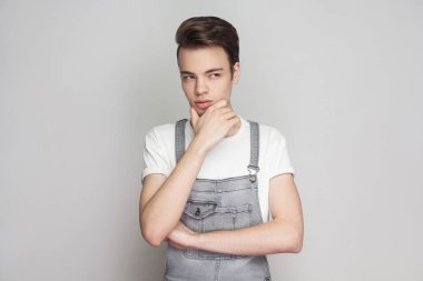 Düşünceli esmer delikanlı ayakta ve uzak gri arka plan üzerinde çene dokunmadan süre seyir beyaz t-shirt ve kot tulum ile rahat tarzı portresi.