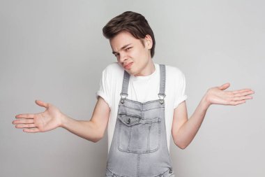 Şüpheli esmer delikanlı eller yayılan ve kameraya gri arka plan üzerinde seyir ile ayakta beyaz t-shirt ve kot tulum ile rahat tarzı portresi