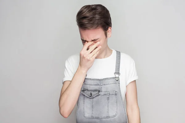 シャツとデニムのオーバー オールを頭を押し 灰色の背景で泣いているカジュアルなスタイルで不幸な失恋若いブルネットの男の肖像 — ストック写真
