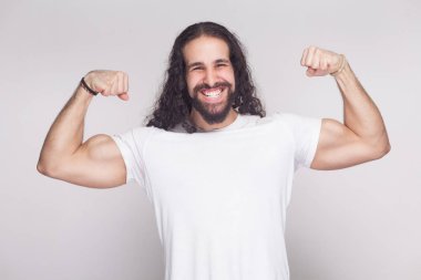 Portrait of strong bodybuilder man in white t-shirt with long wavy hair and beard posing with raised arms and showing biceps with muscular body on gray background  clipart