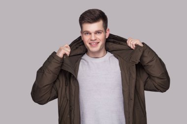 Işık gri gömlek ve kameraya dişlek gülümseme ile seyir gri arka plan üzerinde koyu yeşil parka genç mutlu yakışıklı adam portresi