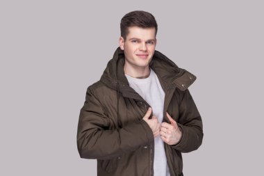 Işık gri gömlek ve kameraya gülümsemek ve memnun yüzü ile seyir gri arka plan üzerinde koyu yeşil parka genç yakışıklı adam portresi