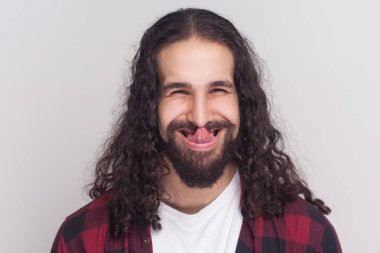 Funny crazy brunette man with beard and black long curly hair in checkered red shirt looking at camera and licking nose on grey background clipart