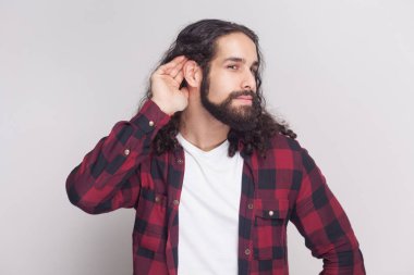 Portrait of nosy man with beard and black long curly hair in checkered red shirt holding hand near ear and listening attentive on grey background clipart