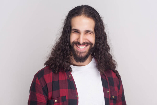 Funny handsome man with beard and black long curly hair in checkered red shirt  winking and looking at camera with toothy smile on grey background