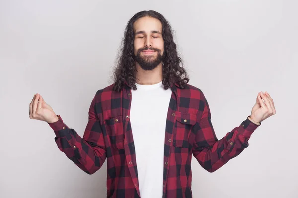 stock image Portrait of calm meditative handsome man with beard and black long curly hair in checkered red shirt standing with closed eyes and showing yoga gesture on grey background 