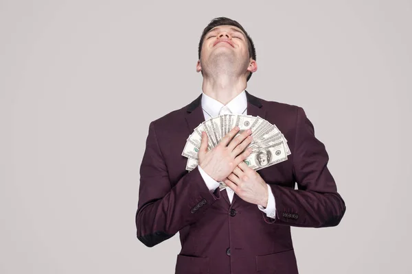 satisfied young businessman in classic violet coat with closed eyes leaning fan of money to chest on gray background