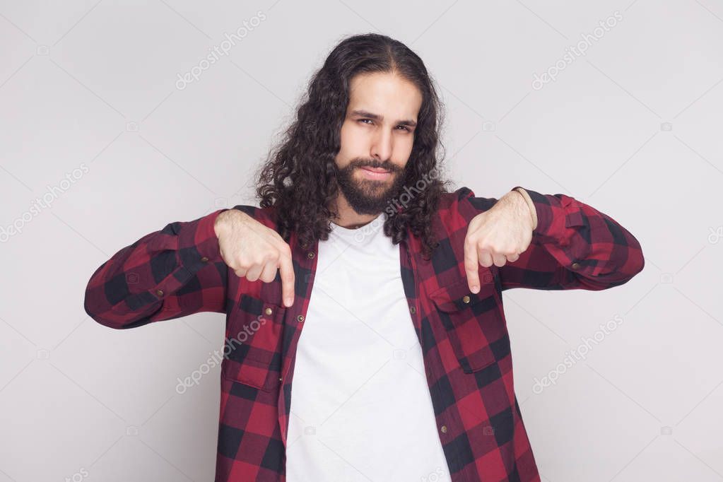 serious handsome man with beard and black long curly hair in checkered red shirt looking at camera and pointing down while showing right now gesture on gray background