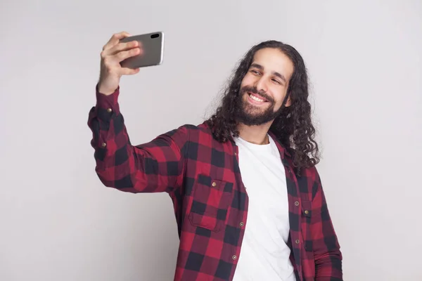 年轻的成人男子在红色格子衬衫和黑色长卷发站立和制作自拍与智能手机在灰色背景 — 图库照片